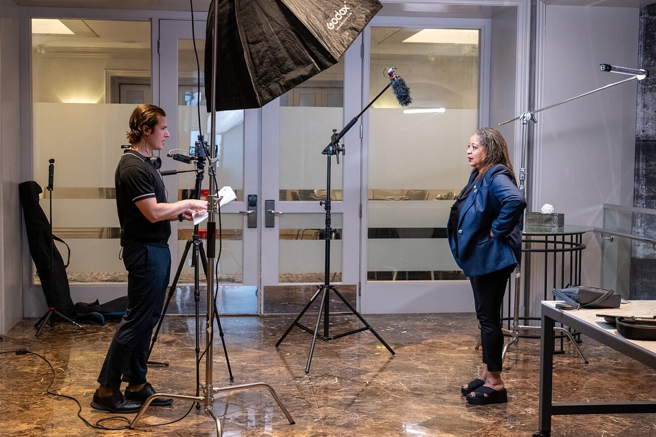 Film crew interview setup, camera on a tripod and overhead lighting present.