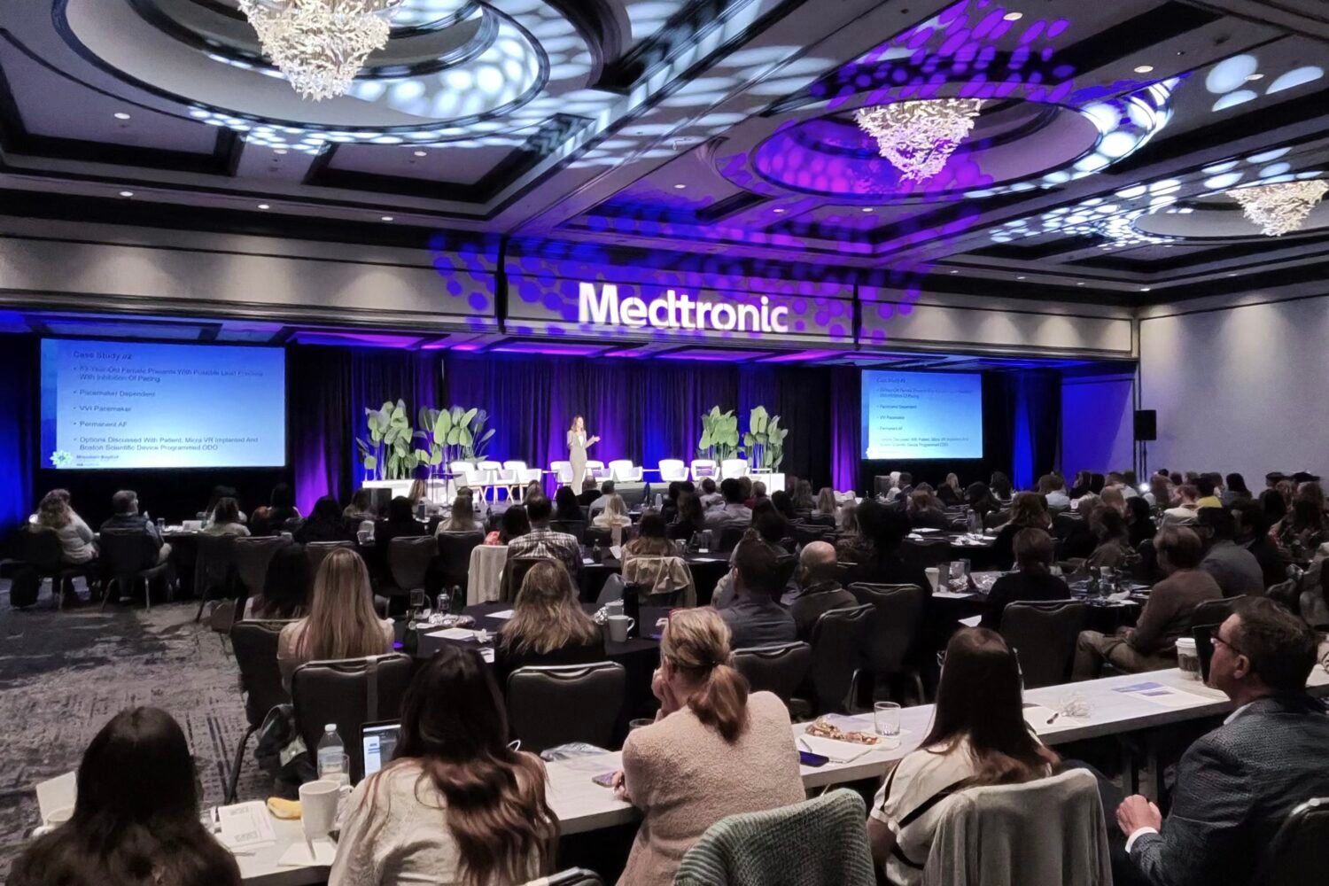 Conference hall with audience seated at tables facing a stage; a purple-lit Medtronic sign on the backdrop.