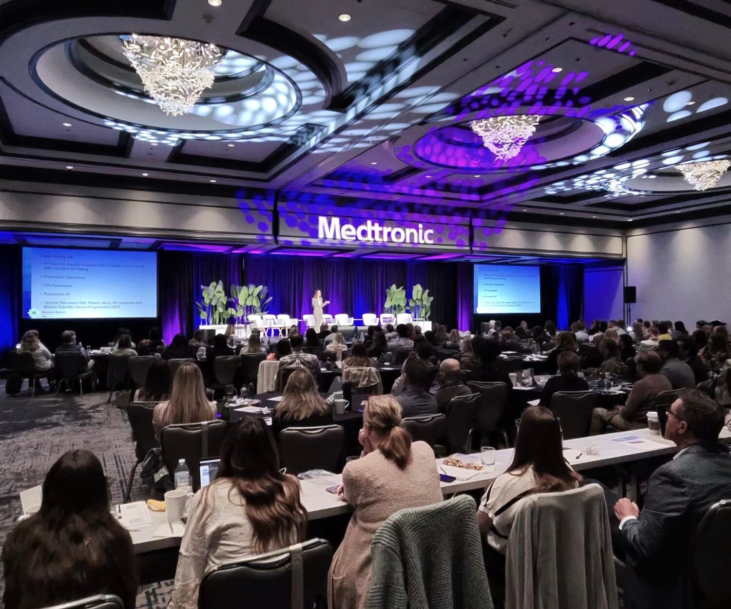 Conference hall with audience seated at tables facing a stage; a purple-lit Medtronic sign on the backdrop.