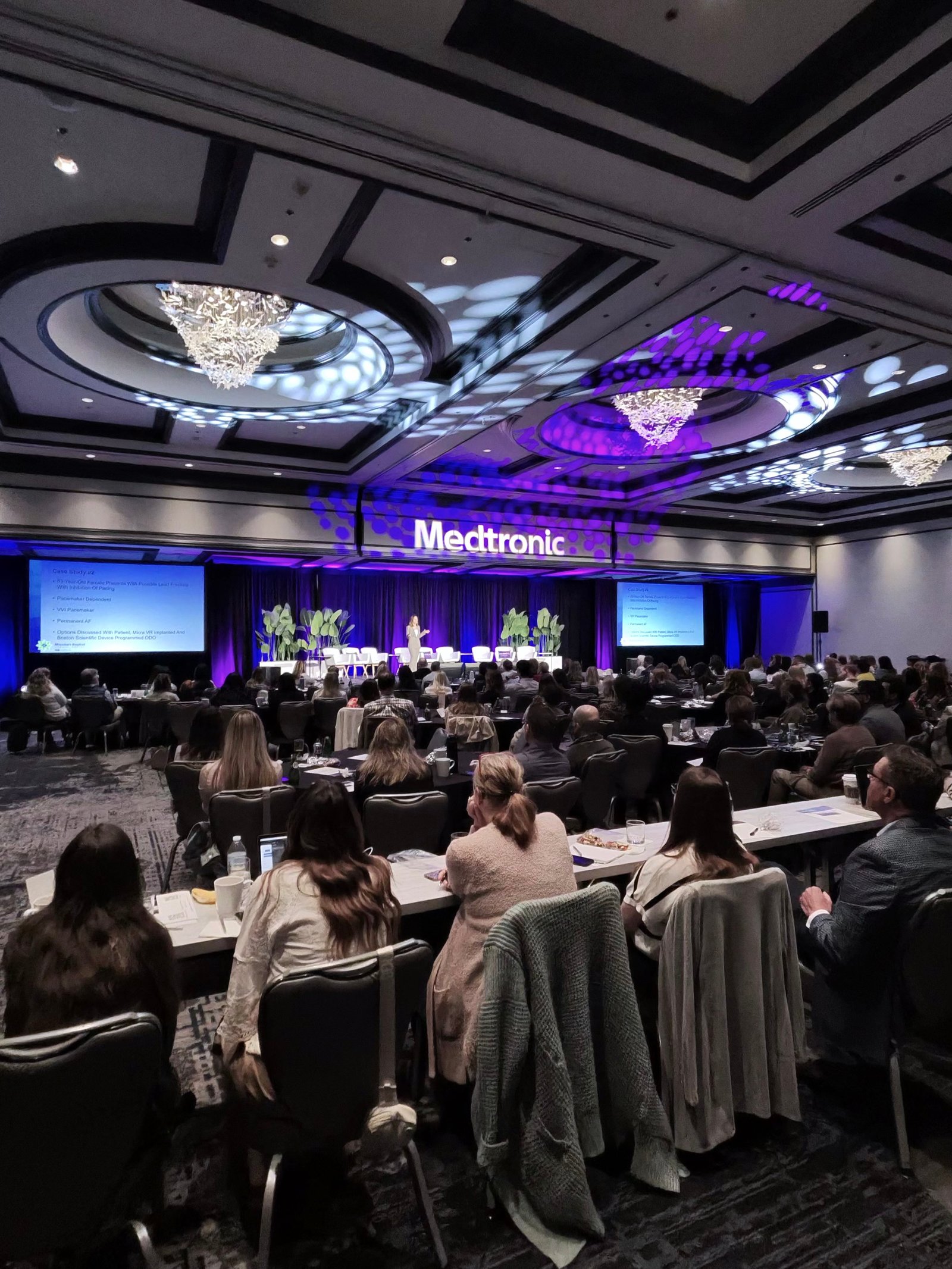 Conference hall with audience seated at tables facing a stage; a purple-lit Medtronic sign on the backdrop.
